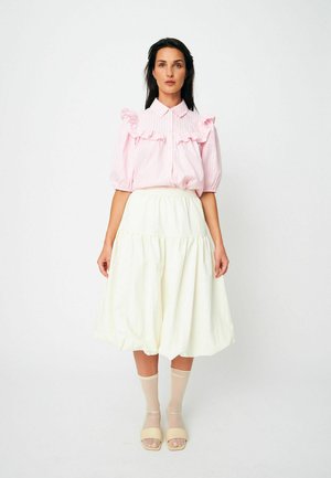 Kobieta stojąca, ubrana w różową bluzkę z falbankami, białą midi spódnicę, przezroczyste beżowe skarpetki oraz beżowe sandały na odkryte palce na jednolitym białym tle.