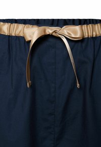 Sötétkék vászon szoknya rugalmas derékpánttal és fényes arany szaténszalagos megkötővel. Finom textúrával és minimális varrási részletekkel rendelkezik.