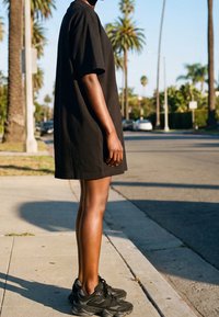 Personne portant une robe t-shirt noire oversized et des baskets noires, debout sur un trottoir en banlieue ensoleillée avec des palmiers en arrière-plan.