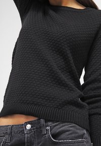 Svart texturerad tröja med bikakestruktur, kombinerad med blekta svarta jeans, som visar en del av magen och en upplyft arm.