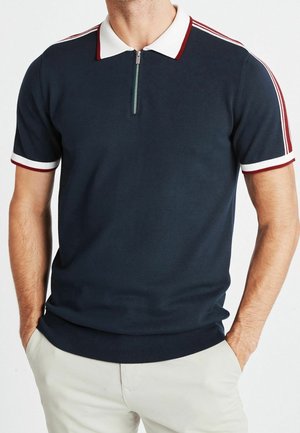 Polo bleu marine avec des rayures rouges et blanches sur le col et les manches. Il dispose d'une courte fermeture éclair au niveau du cou. Fabriqué en tissu lisse.