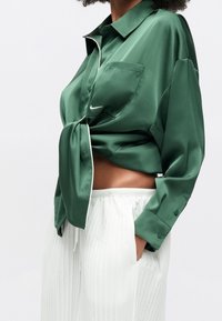 Personne portant une chemise en satin verte boutonnée nouée à la taille avec un pantalon blanc plissé, main dans la poche, vue du cou aux hanches.