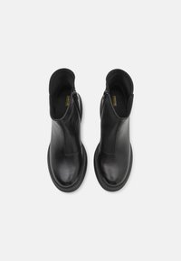 Botas de tobillo de cuero negro con un acabado suave, que cuentan con una cremallera lateral y una puntera redondeada, situadas sobre una suela negra minimalista.