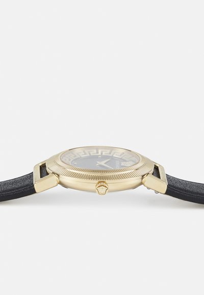 Reloj dorado con correa de piel negra, esfera redonda con un patrón geométrico y un bisel texturizado. Corona detallada con una cabeza de león.