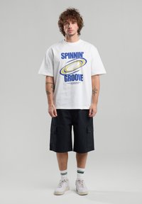 T-shirt en coton blanc avec un texte graphique bleu et jaune "SPINNIN' GROOVE". Associé à un short cargo noir et des baskets blanches avec des accents verts.