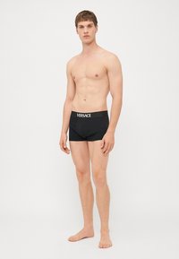 Svarte boxer shorts med en tettsittende design og Versace-logo på livet. Glatt stoff med lett glans, brukt av en stående mannlig modell.