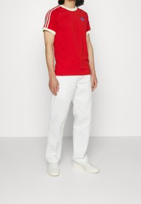 Červené tričko s bílými detaily a modrým logem, zkombinované s bílými kalhotami a světlými teniskami. Krátké rukávy a kulatý výstřih.