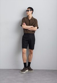 Cămașă scurtă maro cu mâneci scurte, pantaloni scurți negri, șosete negre, mocasini maro cu tălpic alb. Modelul stă în fața unui zid gri.