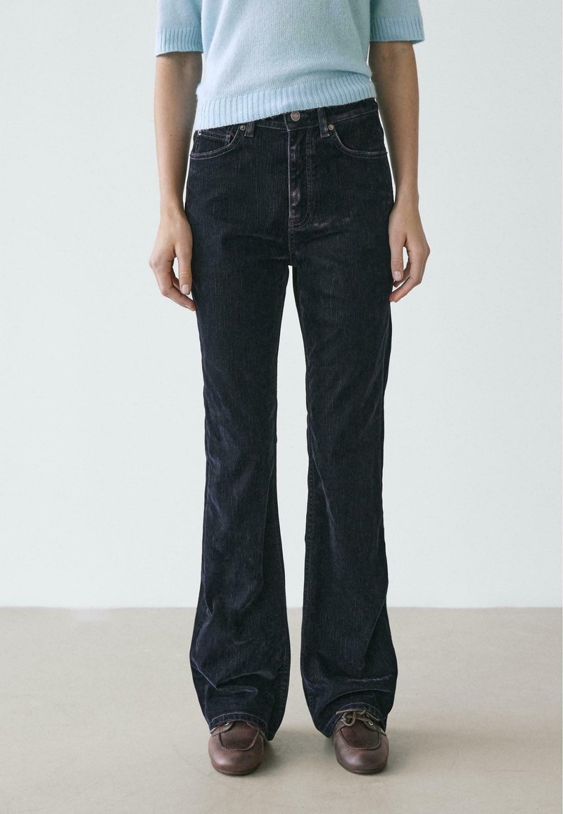 Jeans a zampa in velluto a coste nero con vita alta, dotati di cinque tasche e cuciture chiare. Abbinati a un maglione azzurro chiaro.