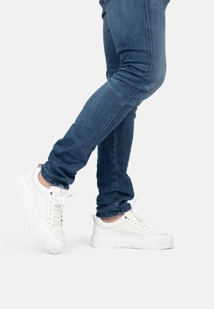 Des sneakers blancs avec un matériau texturé, une semelle plate et un bord surélevé, associés à un jean bleu bien ajusté. Le pantalon présente un détail de couture sur le côté.