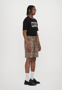 Zwart t-shirt met metallic grafiek, panterprint shorts, zwarte instappers, witte crew-sokken, staande pose, simpele witte achtergrond.