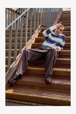 Knit sweater in white and blue stripes, brown wide-leg pants, and brown shoes, seated on rust-colored metal stairs with a railing.