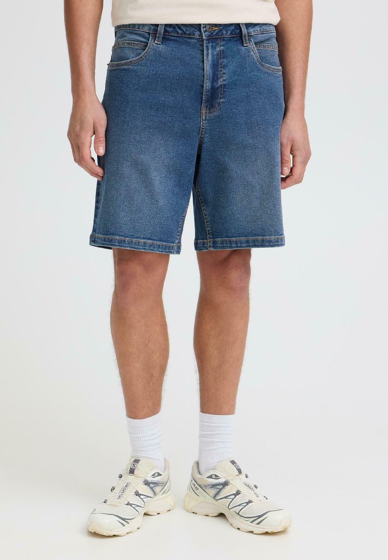 Homme portant un short en denim bleu mi-long, des chaussettes blanches montantes et des chaussures de sport blanches, debout sur un fond blanc.