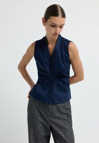 Blusa senza maniche blu navy con scollatura a V, caratterizzata da pieghe in vita e chiusura anteriore con bottoni, realizzata in tessuto liscio.