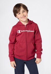 Lächelndes Kind in einem roten Zip-Hoodie mit "Champion"-Logo und blauen Hosen, das vor einem schlichten weißen Hintergrund steht.
