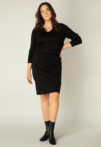 Zwarte, aansluitende jurk met een v-hals en lange mouwen, gemaakt van zacht materiaal. Gelaat met zwarte enkellange laarzen. Eenvoudig, elegant ontwerp.