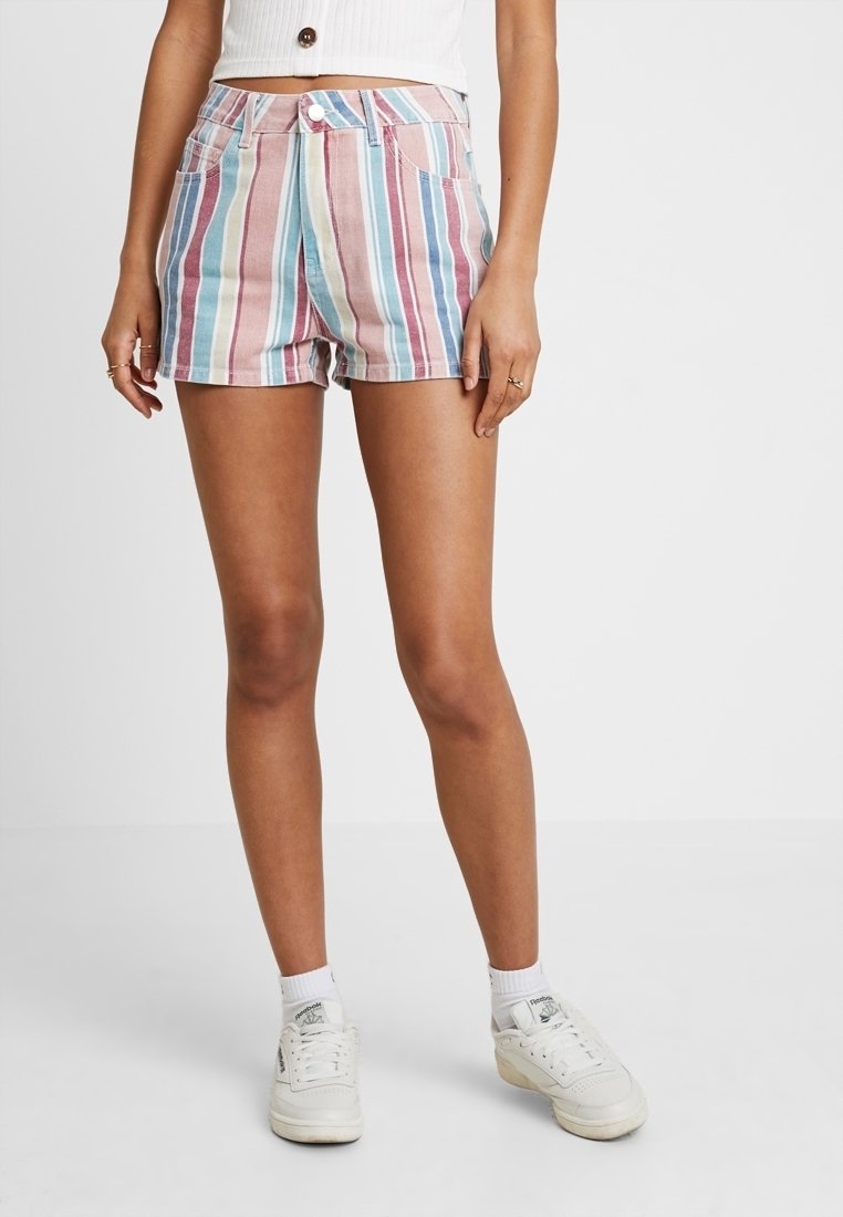 Femme portant un short rayé vertical multicolore, un top court blanc, des chaussettes blanches et des baskets blanches, debout devant un fond uni.