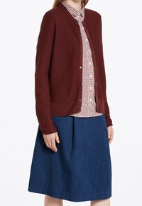Cardigan bordeaux con scollo rotondo e bottoni frontali, indossato sopra una camicia a quadri con colletto e una gonna svasata blu scuro.