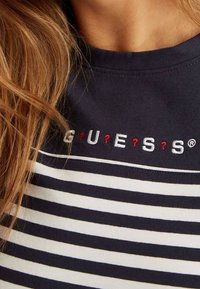 Zbliżenie na niebiesko-białą koszulę w paski z wyszytym napisem "GUESS" oraz czerwonymi znakami zapytania na klatce piersiowej, noszoną przez osobę o jasnobrązowych włosach.