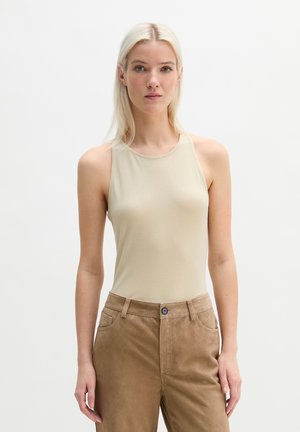 Donna bionda in piedi, che indossa un top beige senza maniche e pantaloni marrone chiaro a vita alta, su uno sfondo bianco semplice.