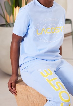 Kind draagt lichtblauw Lacoste T-shirt en broek met geel logo, zittend op een houten blok binnen.
