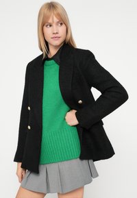 Črn strukturiran blazer z zlatimi gumbi, prekriven z zelenim rebrastim puloverjem in sivimi naborkami.