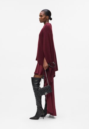 Burgundy stickad poncho över en bordeaux klänning, tillsammans med åtsittande svarta stövlar som går upp över låren och en liten svart quiltad väska med silvernitar.