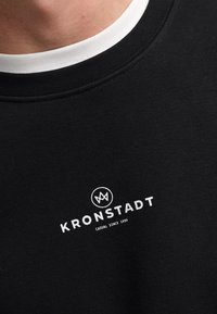 Sudadera negra con cuello redondo blanco, tejido texturizado y un pequeño logotipo en blanco con el texto "KRONSTADT" y "CASUAL SINCE 1999".