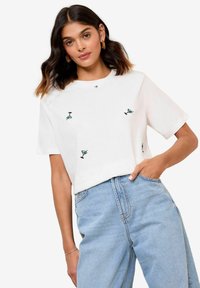 T-shirt en coton blanc à manches courtes, ornée de petits verres à martini brodés en turquoise et noir, portée avec un jean bleu taille haute.