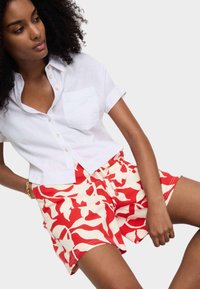 Weißes Leinenhemd mit kurzen Ärmeln und Brusttasche, kombiniert mit rot- und cremefarbigen Shorts mit abstrakten Mustern.