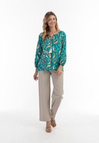 Blusa turchese a maniche lunghe con motivo paisley e collo con coulisse, abbinata a pantaloni beige a gamba larga e sandali con cinturino color sabbia.