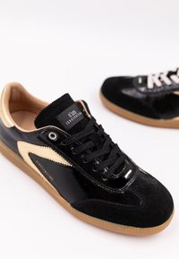 Svarta sneakers i patentläder och mocka med en gummisula. Har en gyllene accent och svarta snören. Logotypen på tungan.