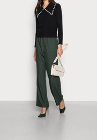 Maglione nero a maglia con un ampio colletto decorato con dettagli floreali bianchi, abbinato a pantaloni larghi verdi e una piccola borsa beige.