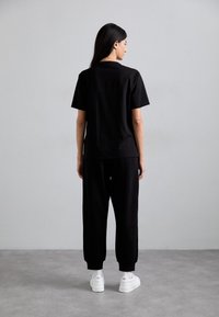 Zwarte katoenen T-shirt met korte mouwen, gecombineerd met zwarte sweatpants. Draagt witte sneakers, van achteren gezien tegen een lichte achtergrond.