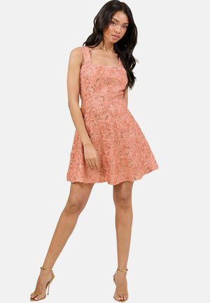 Kurzes pinkfarbenes Kleid mit quadratischem Ausschnitt, strukturierten Rosenmustern, tailliertem Oberteil und ausgestellt Rock. Kombiniert mit goldenen Riemchenschuhen.