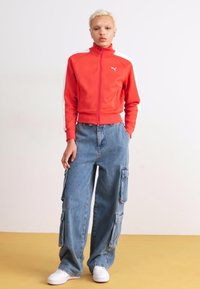Veste zippée rouge avec des rayures blanches, associée à un jean cargo bleu ample avec des poches latérales et des baskets blanches sur un sol jaune.