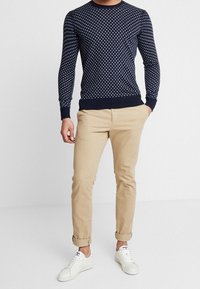 Muž v tmavě modrém svetru s bílými puntíky, béžových nohavicích s ohrnutými lemy a bílých teniskách stojí na jednobarevném bílém pozadí.