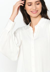 Camicia bianca con bottoni, colletto strutturato, maniche voluminose e una texture liscia. Presenta bottoni perlacei lungo il fronte.