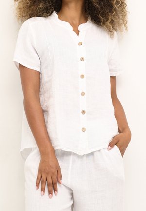 Kvinde med en hvid kortærmet hørskjorte med træknapper og matchende hvide hørbukser, hånd i lomme.