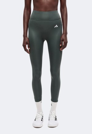 Osoba nosiaca tmavozelené športové legíny s vysokým pásom, biele ponožky a biele tenisky Adidas, stojaca pred jednoduchým pozadím.