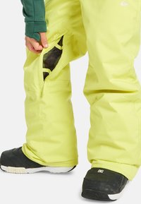 Žlté nepremokavé snehové nohavice s ventiláciou so sieťovinou na zips, nastaviteľný strih a čierna obuv s bielymi akcentmi.