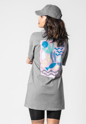 Frau mit welligem Haar, die eine graue Mütze und ein langes graues T-Shirt mit einem bunten abstrakten Naturdesign auf der Rückseite trägt, kombiniert mit schwarzen Shorts.