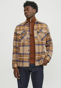 Bruin geruite blouse met blauwe en gele accenten, met twee borstzakken en zwarte knopen. Draagt over een bruine opstaande kraag.