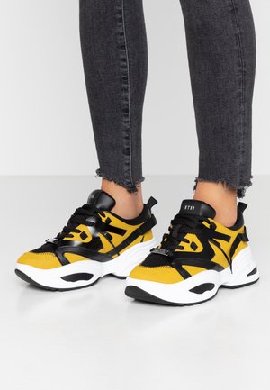 Persoon met gele en zwarte chunky sneakers met dikke witte zolen en zwarte veters, gecombineerd met donkerkleurige skinny jeans met rafelige zoom.