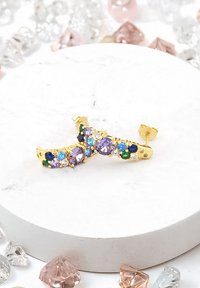 Orecchini color oro con gemme colorate—viola, verde, blu—impostati in un design curvo, appoggiati su una superficie in marmo circondata da cristalli.