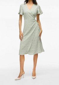 Kvinna iklädd en ljusgrön omslagsklänning med subtilt blommönster och korta ärmar, i kombination med beige högklackade slingbackskor.