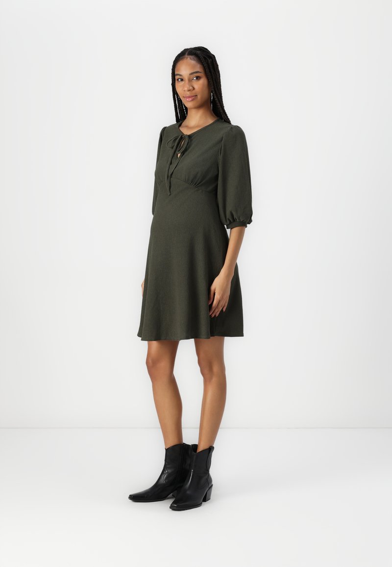 Olivengrønn kjole med rund hals, puffærmer og knytedetalj. Kombinert med svarte ankelstøvler som viser en helårs, utsvingt design.