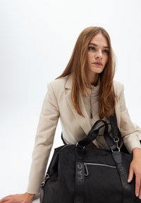 En kvinna med långt brunt hår bär en krämfärgad blazer, sitter med en svart gymväska som vilar bredvid henne, och visar ett eftertänksamt uttryck.