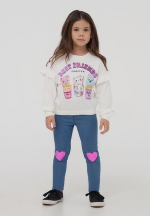 Felpa bianca con dettagli a volant, caratterizzata da grafiche colorate di animali e testo "Amici per sempre". Jeans blu con toppe a forma di cuore rosa.