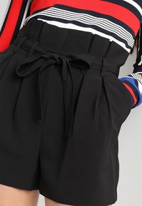 Černé šortky s vysokým pasem, s uvázaným látkovým opaskem, které mají sklady a boční kapsy. Hladká textura s upraveným, strukturovaným designem.